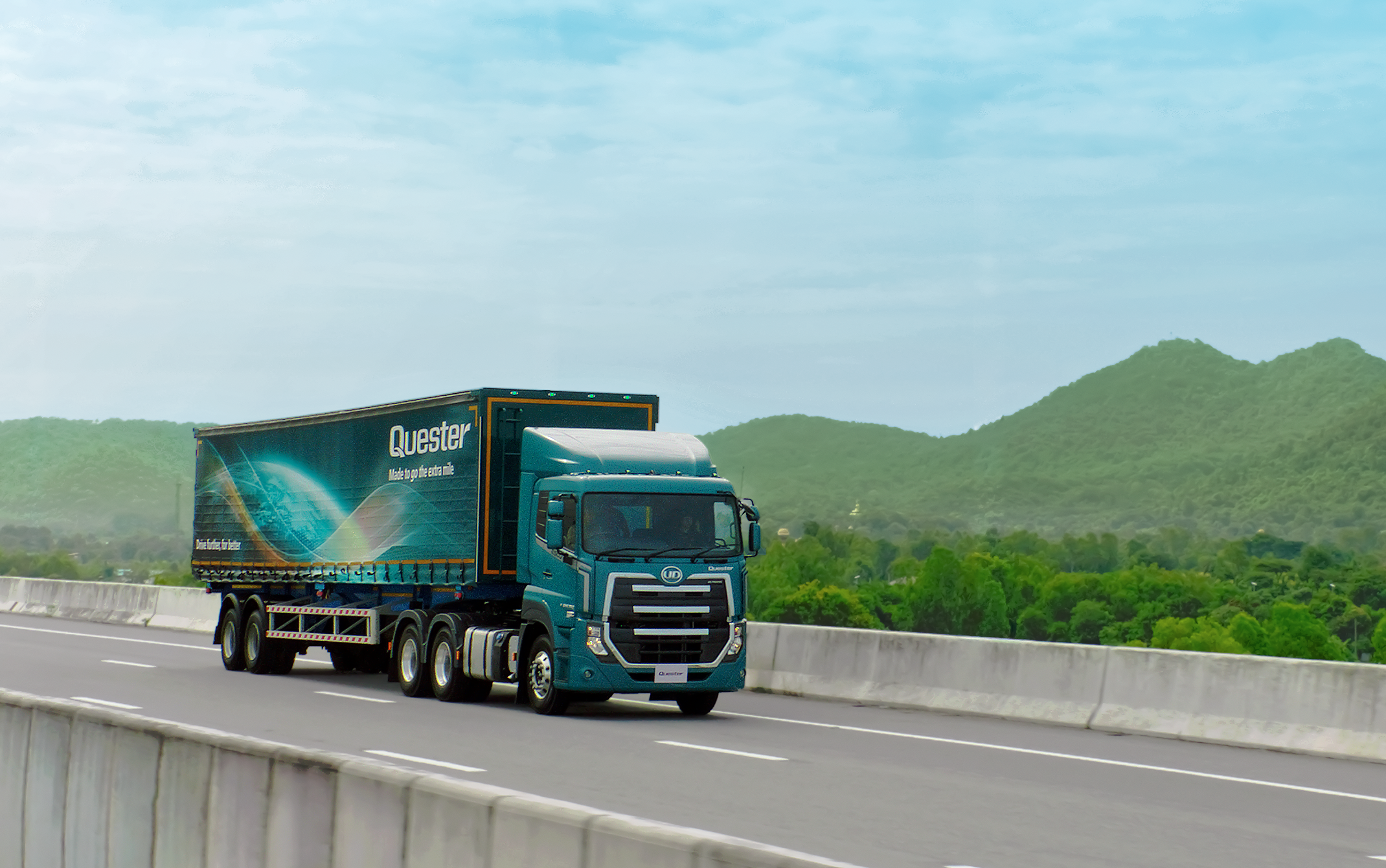 Quester Truck driving on a highway bridge with green hills and blue sky in the background.