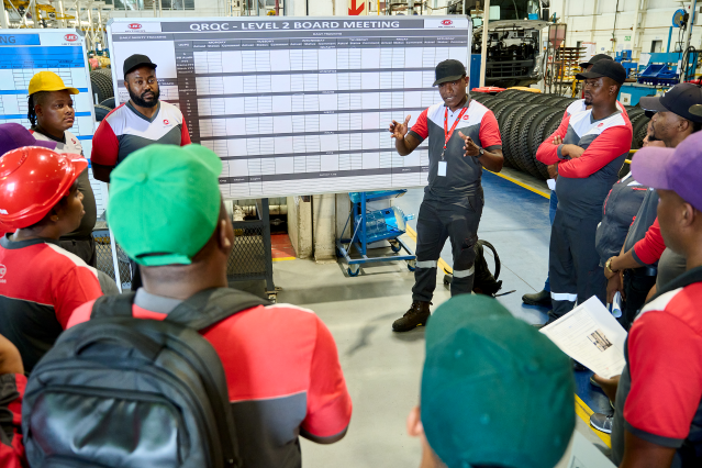 Werknemers in 'n fabrieksvloer het 'n vergadering voor 'n groot witbord.