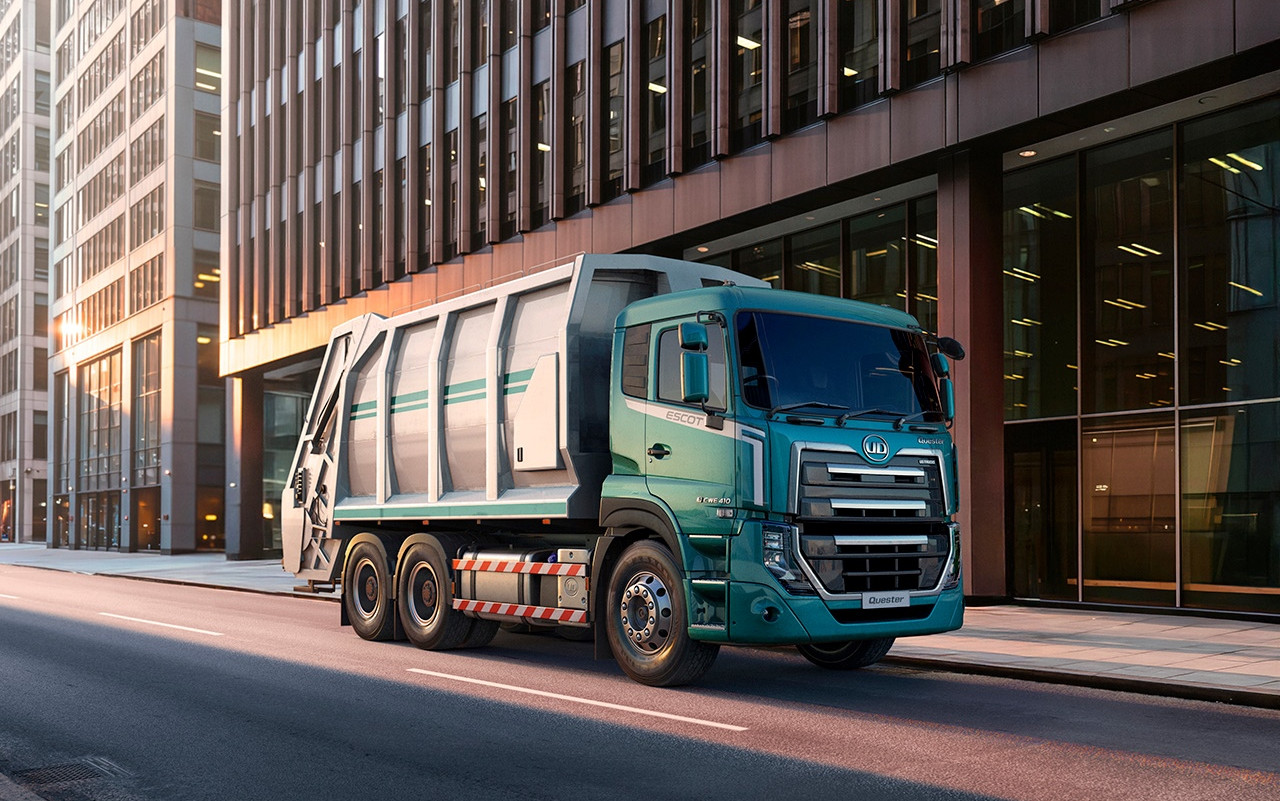 Green garbage truck driving on an empty city street lined with tall modern buildings.