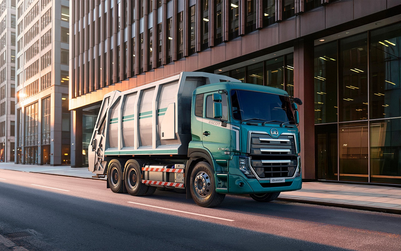 Green garbage truck on empty city street lined with tall glass buildings at sunrise.