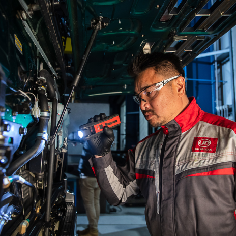 Mechanic in uniform inspects machinery with flashlight, wearing safety glasses.